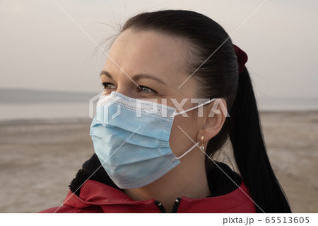 A brunette woman in sunglasses and a red jacket and a protective surgical mask on an empty beach during quarantine. 65513605
