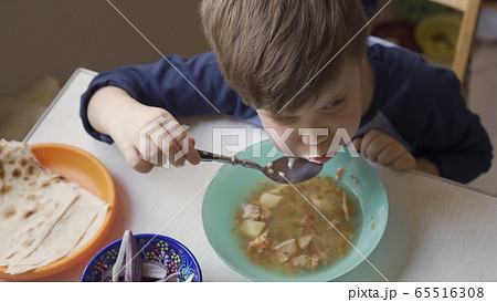 Boy eats soup sitting at dining room table with fresh onion and pita bread on it. High angle view 65516308