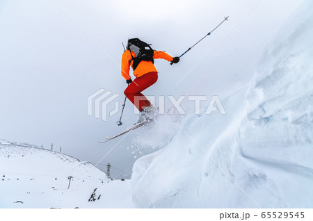 Male athlete skier in an orange trigger with a gray backpack and ski poles jumps from a high snowy slope against the backdrop of mountains in the clouds on a cloudy day. Male athlete skier in an orange trigger with a gray backpack and ski poles jumps from a high snowy slope against the backdrop of mountains in the clouds on a cloudy day. 65529545
