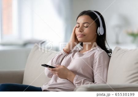 African woman wears headphones listen classic music sitting on couch 65530199