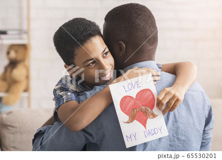 Little afro boy hugging dad and giving him handmade postcard Little afro boy hugging dad and giving him handmade postcard 65530201
