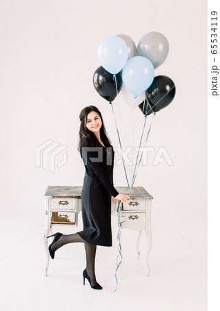 glad positive Caucasian girl in black dress holds bunch of air balloons, standing near vintage white table with drawers, waits for guests, isolated over white background. Birthday party concept 65534119