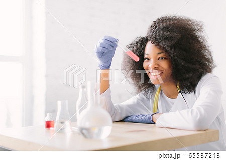 Young beautiful African American girl doctor in a white coat with a stethoscope. sitting at a table with reagent flasks on white background. Female lab assistant studying blood sample for analysis 65537243