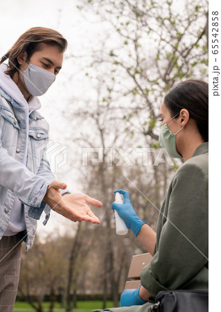 Young man in mask opening palms while his girlfriend spraying sanitizer Young man in mask opening palms while his girlfriend spraying sanitizer 65542858