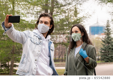 Young man and his girlfriend in casualwear and protective masks making selfie 65542880
