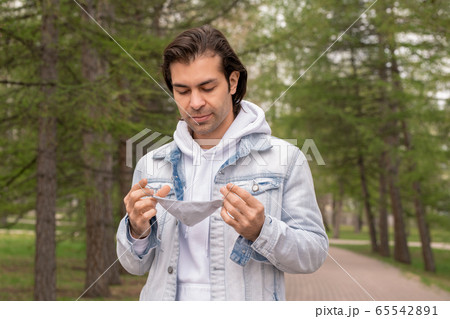 Young contemporary man in hoodie and denim jacket holding protective mask Young contemporary man in hoodie and denim jacket holding protective mask 65542891