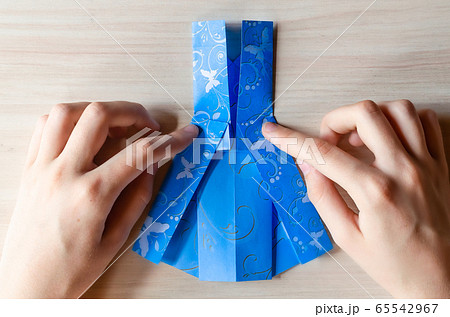 Girl's hands making origami blue dress from butterfly printed paper. Step by step instruction, step16. Mother's day, birhday, summer gift, family leisure time concept. DIY tutorial, flatlay, top view 65542967