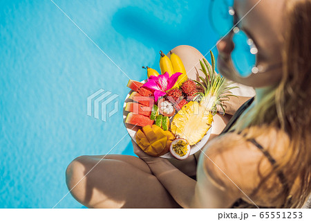 Young woman relaxing and eating fruit plate by the hotel pool. Exotic summer diet. Photo of legs 65551253