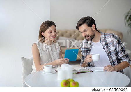 Cute young woman in a white dress holding a tablet in her hand and showing something to her husband Cute young woman in a white dress holding a tablet in her hand and showing something to her husband 65569095
