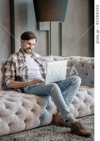 Dark-haired bearded man on the couch with a laptop. Dark-haired bearded man on the couch with a laptop. 65569303