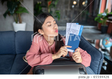 Happy dark-haired female sitting on sofa, holding airplane tickets, her lips giving kiss 65594426