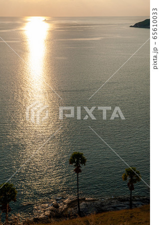 View on Palm tree and islands near sea water during beautiful sunset with a bright yellow sun shining rays over the rocks. Suited for a wallpaper background with a soft yellow sky. Sun over water. 65610033
