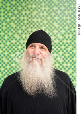 Mature bearded hipster man with beanie hat thinking against green wall Mature bearded hipster man with beanie hat thinking against green wall 65616351