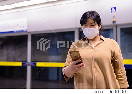 Overweight Asian woman using phone with mask for protection from corona virus outbreak at subway 65616635