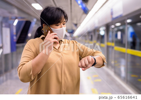 Overweight Asian woman with mask talking on the phone and checking time at subway train station 65616641