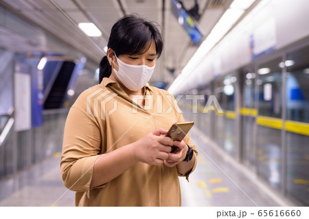 Overweight Asian woman using phone with mask for protection from corona virus outbreak at subway 65616660