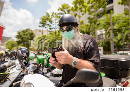 Mature bearded hipster man with sunglasses and mask using phone while sitting on motorcycle Mature bearded hipster man with sunglasses and mask using phone while sitting on motorcycle 65616984