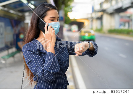 Young Asian businesswoman with mask checking smartwatch and talking on the phone at bus stop 65617244