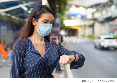 Young Asian businesswoman with mask checking smartwatch and waiting at the bus stop Young Asian businesswoman with mask checking smartwatch and waiting at the bus stop 65617245