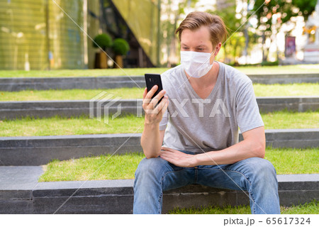Young man with mask for protection from corona virus outbreak using phone while sitting outdoors 65617324