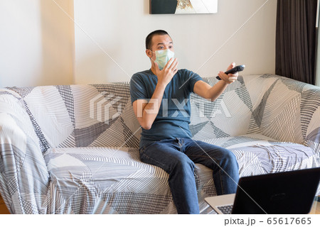 Young Asian man with mask watching tv and looking shocked at home under quarantine 65617665