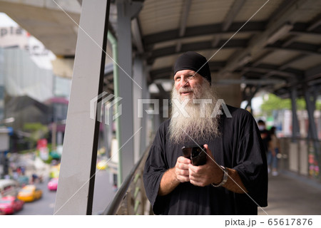 Happy mature handsome bearded hipster man thinking while using phone at footbridge in the city 65617876