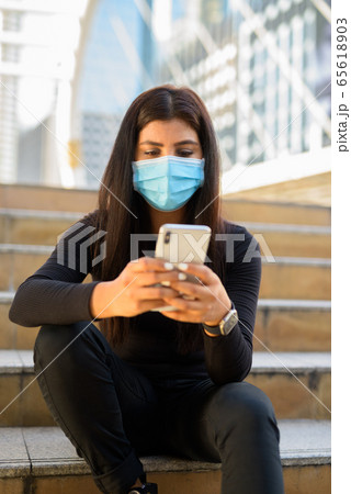 Young Indian woman with mask using phone and sitting by the stairs in city Young Indian woman with mask using phone and sitting by the stairs in city 65618903