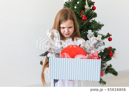 Shocked little child girl looks with opened eyes and worried expression, holding box with various plastic wastes over christmas tree background. Shocked little child girl looks with opened eyes and worried expression, holding box with various plastic wastes over christmas tree background. 65638808