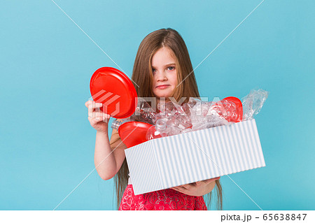 Shocked little child girl looks with opened eyes and worried expression, holding box with various plastic wastes on blue background. 65638847