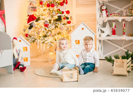 Cute little boy with blond hair plays with little girl in a bright room decorated with Christmas garlands near the Christmas tree. Happy childhood. Christmas mood Cute little boy with blond hair plays with little girl in a bright room decorated with Christmas garlands near the Christmas tree. Happy childhood. Christmas mood 65639077
