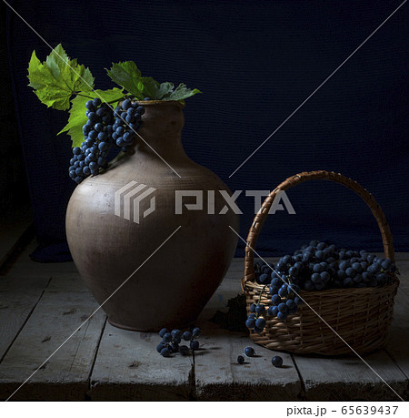 Still life with a big old jug and wild grapes. A bunch of blue grapes with bright green leaves lies in a jug. A lot of grapes lies in a wicker basket next to a jug on a wooden table. Country style. Still life with a big old jug and wild grapes. A bunch of blue grapes with bright green leaves lies in a jug. A lot of grapes lies in a wicker basket next to a jug on a wooden table. Country style. 65639437