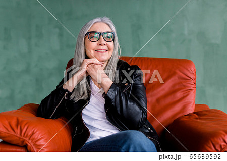 Beautiful stylish senior woman with long straight gray hair, wearing eyeglasses and black leather jacket, sitting in comfortable sofr red armchair on green background and dreamly looking aside Beautiful stylish senior woman with long straight gray hair, wearing eyeglasses and black leather jacket, sitting in comfortable sofr red armchair on green background and dreamly looking aside 65639592