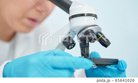Close-up of woman lab technician in blue rubber gloves looking through microscope. 65641026