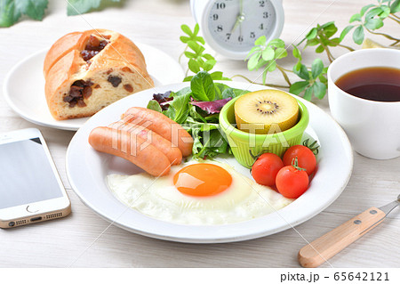 ワンプレート朝食(洋食、目玉焼き、ソーセージ)のイメージ。レーズンパンとコーヒーとスマホと時計。 ワンプレート朝食(洋食、目玉焼き、ソーセージ)のイメージ。レーズンパンとコーヒーとスマホと時計。 65642121