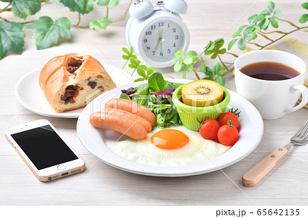 ワンプレート朝食（洋食、目玉焼き、ソーセージ）のイメージ。レーズンパンとコーヒーとスマホと時計。 65642135