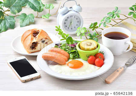 ワンプレート朝食(洋食、目玉焼き、ソーセージ)のイメージ。レーズンパンとコーヒーとスマホと時計。 ワンプレート朝食(洋食、目玉焼き、ソーセージ)のイメージ。レーズンパンとコーヒーとスマホと時計。 65642136