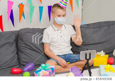 Boy in medicine face mask and festive cap celebrates birthday by video call to phone Boy in medicine face mask and festive cap celebrates birthday by video call to phone 65649366