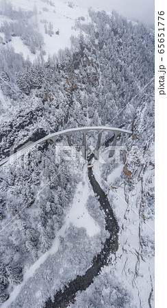 Vertical Aerial panorama of the Landwasser Viaduct with Railway without famous train at winter, landmark of Switzerland, snowing, river and mountains 65651776