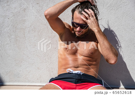 The handsome brutal man in sunglasses with a long hair and naked torso sits near the pool, collects hair in a tail, a sports suntanned body, sunglasses with a blue frame, red blue swimming shorts The handsome brutal man in sunglasses with a long hair and naked torso sits near the pool, collects hair in a tail, a sports suntanned body, sunglasses with a blue frame, red blue swimming shorts 65652618