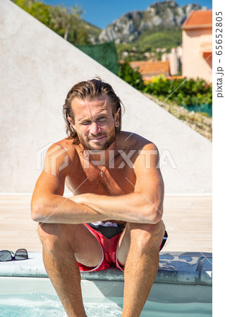 The handsome brutal man in sunglasses with a long hair and naked torso sits near the pool, is looking a camera, a sports suntanned body, sunglasses with a blue frame, he is red blue swimming shorts 65652805
