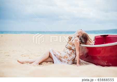 A young girl sits on the beach leaning against a boat 65668555