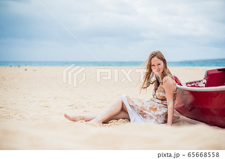 A young girl sits on the beach leaning against a boat 65668558