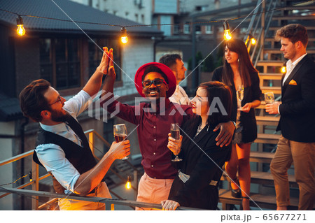Corporate celebrating in the warm light of the lamps in summer evening, young friends, colleagues look happy, talk, have fun Corporate celebrating in the warm light of the lamps in summer evening, young friends, colleagues look happy, talk, have fun 65677271