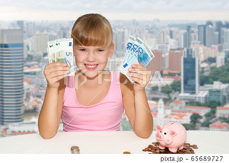 Little girl sitting on the desk with coins and pink piggy bank and holding in hands european currency  65689727