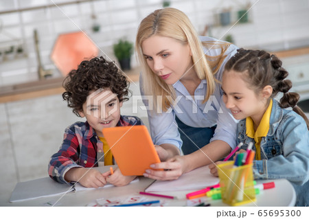 Curly boy and dark-haired girl sitting at table, blonde female bending between them, showing something on tablet 65695300