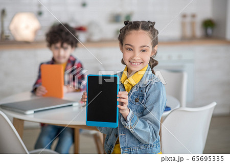 Dark-haired girl showing laptop into the camera, smiling, curly boy sitting at round table with tablet behind her Dark-haired girl showing laptop into the camera, smiling, curly boy sitting at round table with tablet behind her 65695335