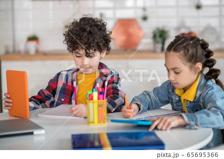Boy and girl sitting at round table, holding tablets, making notes 65695366