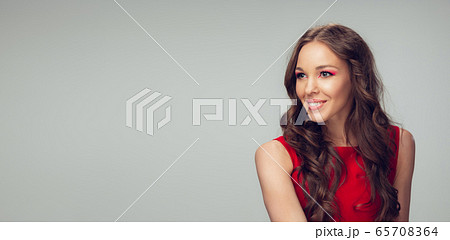 Beautiful young woman with long healthy curly hair and bright make up wearing red dress isolated on grey studio backgroud. Flyer with copyspace. 65708364