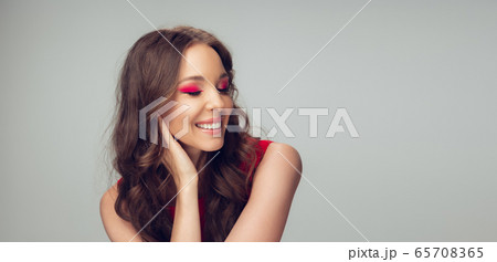Beautiful young woman with long healthy curly hair and bright make up wearing red dress isolated on grey studio backgroud. Flyer with copyspace. 65708365