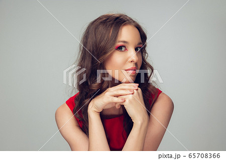 Beautiful young woman with long healthy curly hair and bright make up wearing red dress isolated on grey studio backgroud. Calm, thoughtful. Beautiful young woman with long healthy curly hair and bright make up wearing red dress isolated on grey studio backgroud. Calm, thoughtful. 65708366